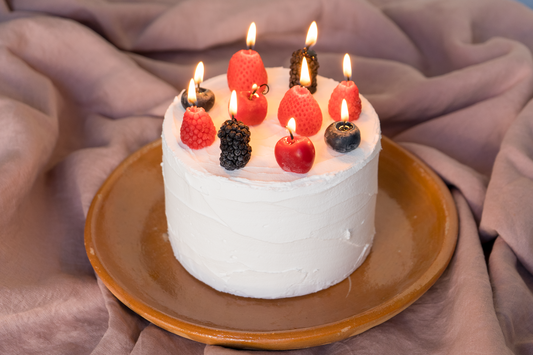 A small white frosted cake topped with lit candles and fresh berries, presented on a round brown plate against a draped mauve fabric backdrop.