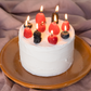 A small white frosted cake topped with lit candles and fresh berries, presented on a round brown plate against a draped mauve fabric backdrop.