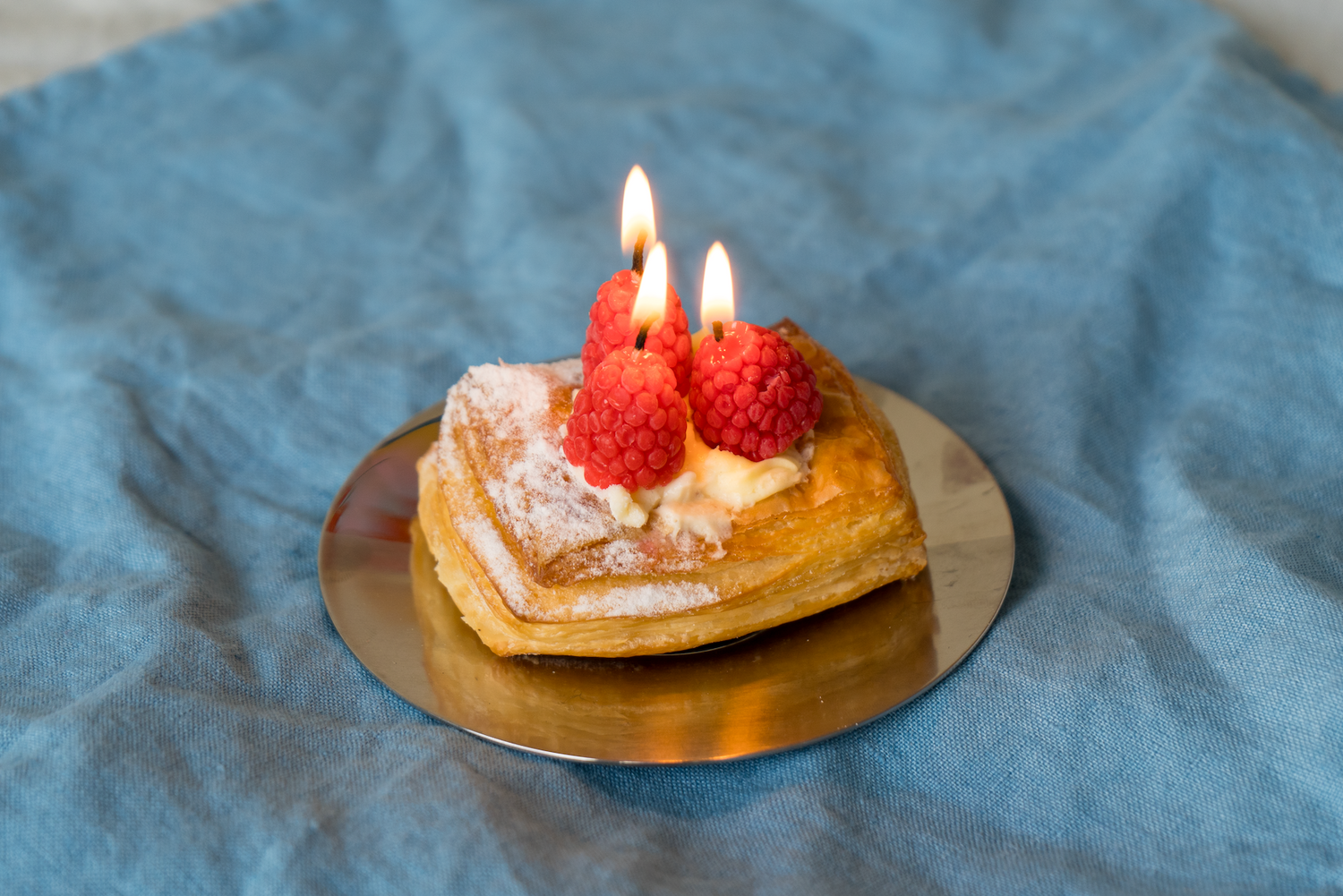 Small pastry with lit candles and raspberry birthday candles on a blue fabric background